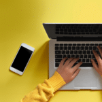 Auf dem Bild ist ein Laptop-Computer zu sehen, auf dem Hände gerade etwas tippen. Daneben steht eine Kaffeetasse, auf der anderen Seite liegt ein Smartphone.