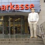 Gebäude mit der Aufschrift Sparkasse. Eine Treppe führt zu dem Gebäude hinauf. Auf der obersten Stufe ist die Statue eines Mannes im Anzug mit den Händen in den Hosentaschen zu sehen.