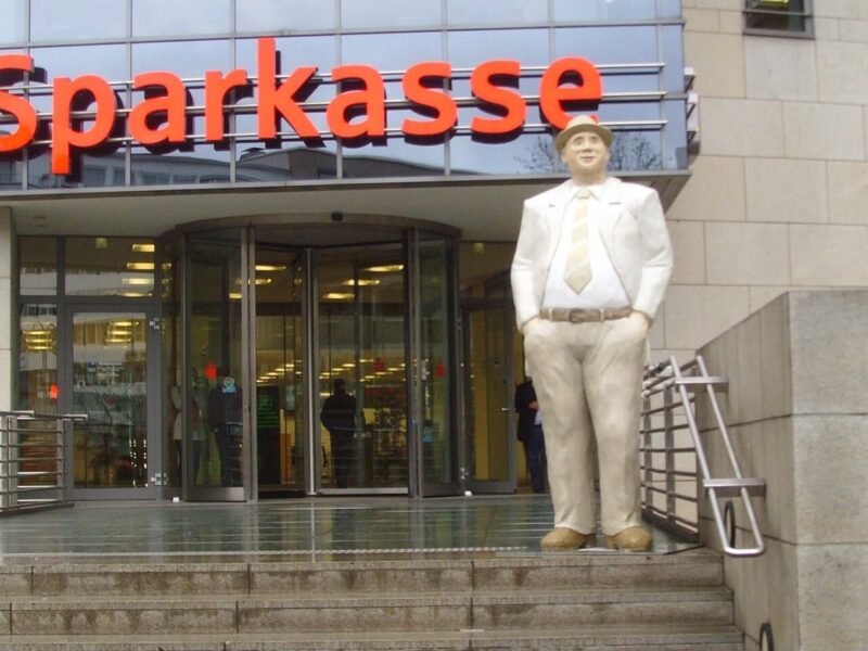 Gebäude mit der Aufschrift Sparkasse. Eine Treppe führt zu dem Gebäude hinauf. Auf der obersten Stufe ist die Statue eines Mannes im Anzug mit den Händen in den Hosentaschen zu sehen.