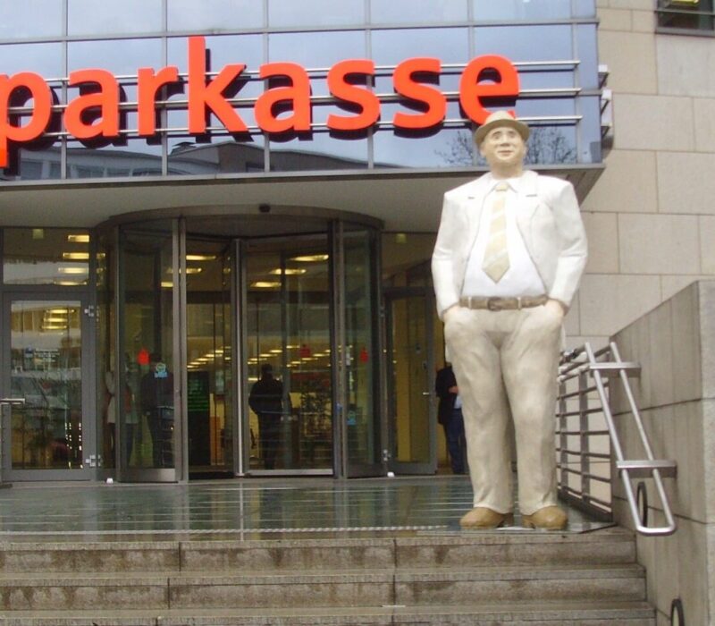 Gebäude mit der Aufschrift Sparkasse. Eine Treppe führt zu dem Gebäude hinauf. Auf der obersten Stufe ist die Statue eines Mannes im Anzug mit den Händen in den Hosentaschen zu sehen.