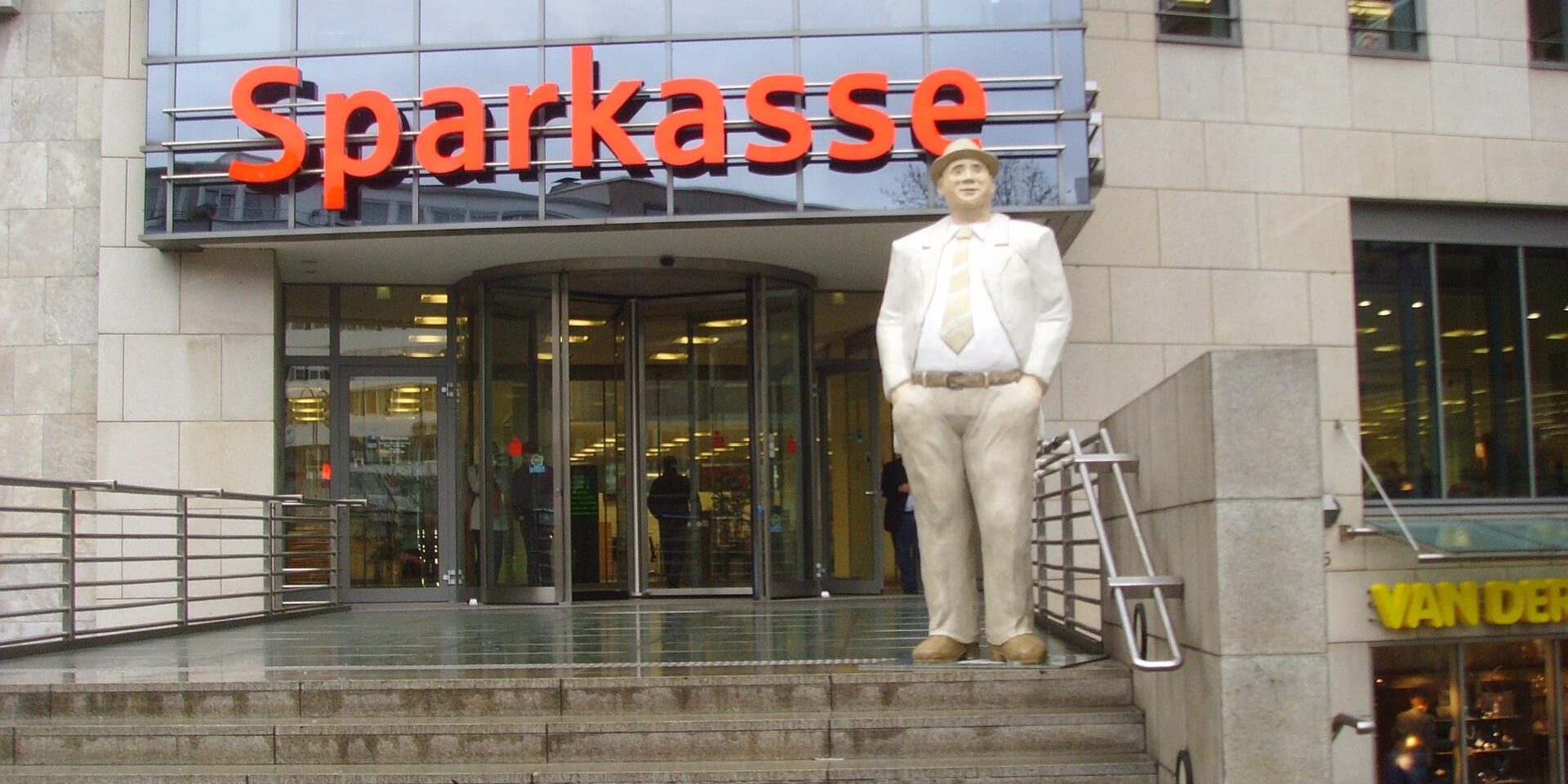 Gebäude mit der Aufschrift Sparkasse. Eine Treppe führt zu dem Gebäude hinauf. Auf der obersten Stufe ist die Statue eines Mannes im Anzug mit den Händen in den Hosentaschen zu sehen.